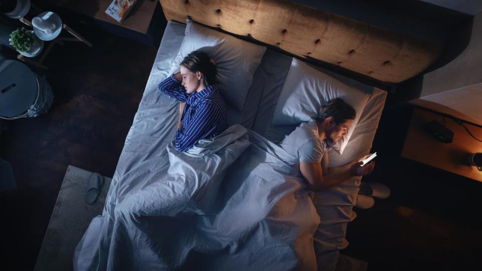 Overhead view of couple laying in bed facing away from each other in the dark while one person is scrolling on phone.