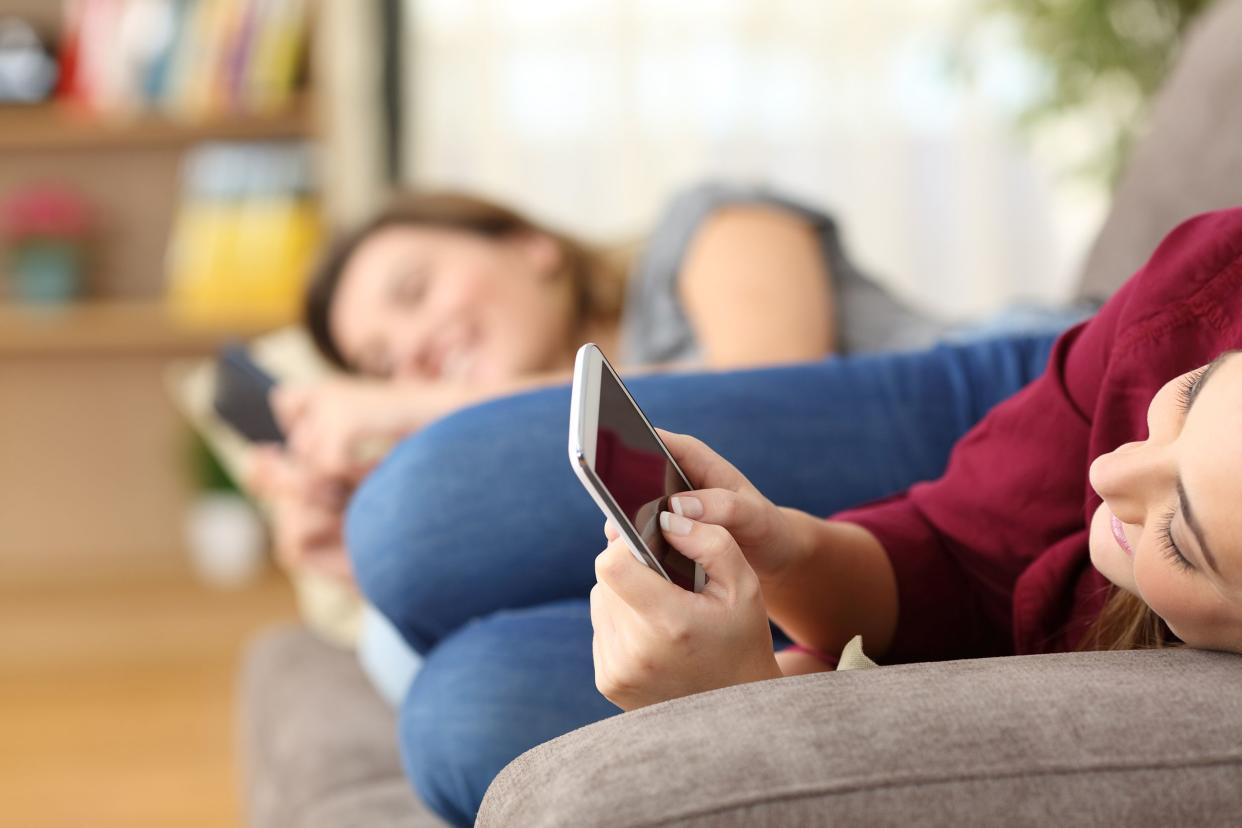 Closeup of side view of one woman on her smartphone while laying on a grey couch, she is looking at the screen, smiling, with a another woman doing the same thing on the other side of the couch and the rest of a vibrant living room blurred in the backgrou