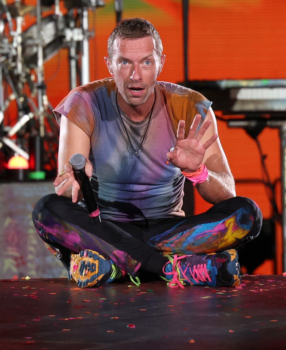 Chris Martin performing on stage, crouched down, gesturing with his hand, in a colorful outfit