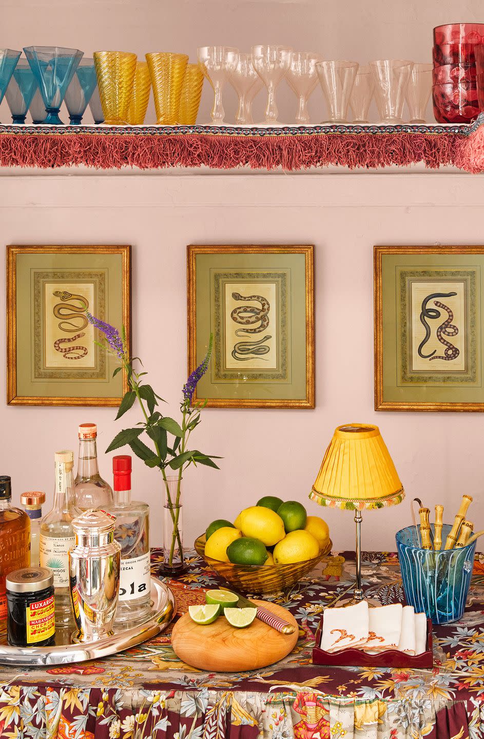 a bar covered in a patterned cloth with a plum colored background has a tray with bottles, a bowl of lemons and limes, and a small lamp, above are three framed snake prints and a shelf with colored glasses