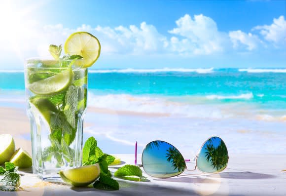 A cocktail drink with fresh lime and mint leaves next to sunglasses on a table, with a beach in the background.