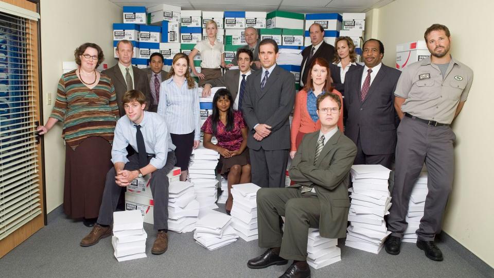 the office us cast group shot, with people either standing or sitting on stacks of paper