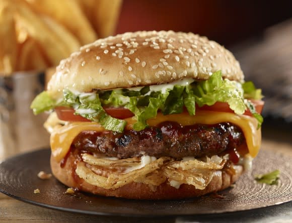 Burger with sesame seed bun, lettuce, tomato, cheese, onions, and BBQ sauce on a plate.