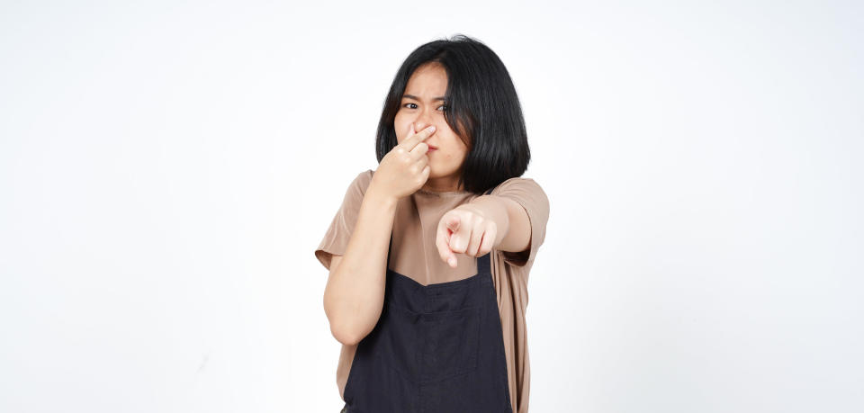 An image of a woman with short black hair in a bob, pointing at the camera with her nose held in her other hand as though she's smelled something bad