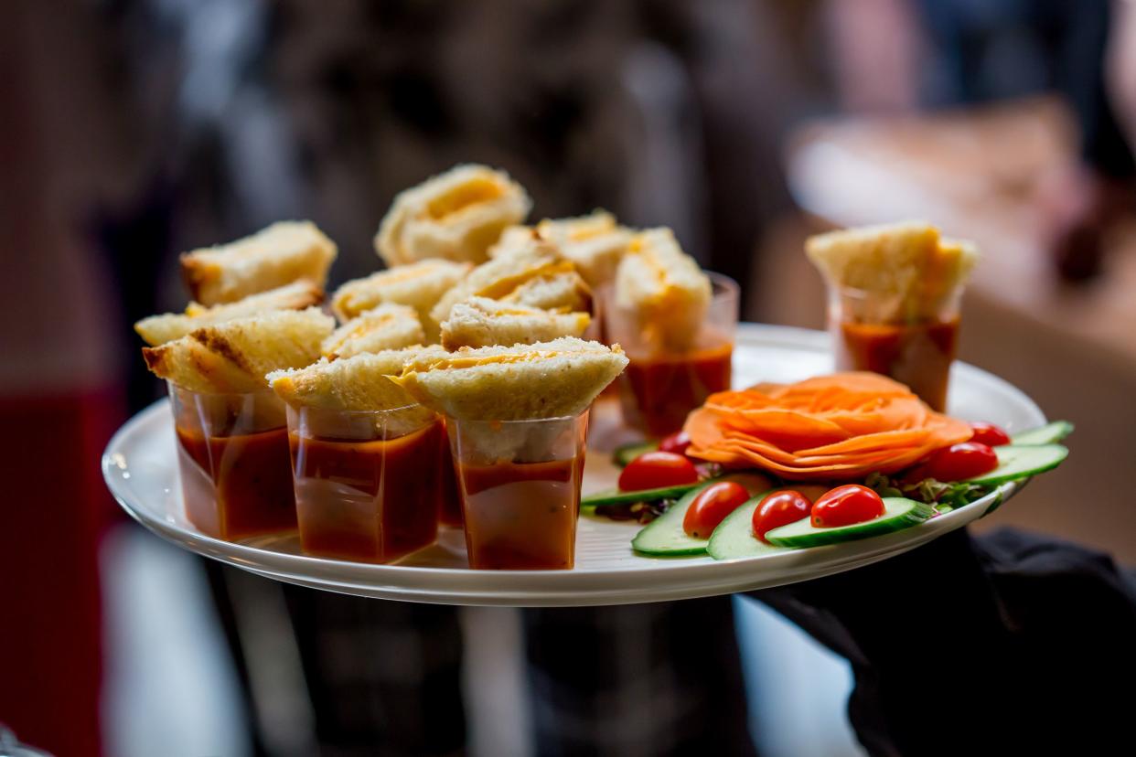 Focus on a metal tray of mini grilled cheese appetizers with a male server holding it with a blurred background of an upscale gathering