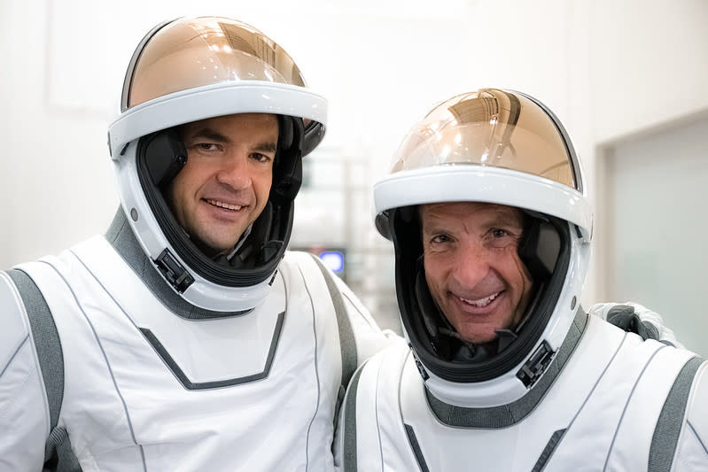 Two men in white spacesuits, seen from the chest up. Their copper-colored visors are raised, revealing their faces inside their helmets. They are in a white room, smiling.