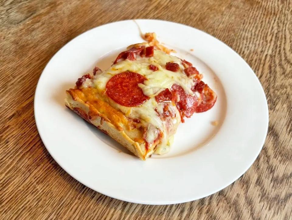 A square-shaped piece of pizza with oozing cheese and pepperoni on a white plate placed on a wooden table