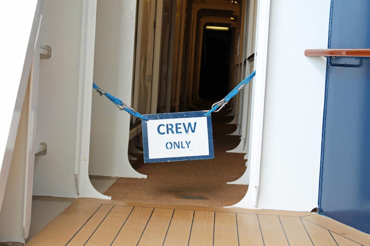 'Crew Only' sign hung in the doorway of a hallway of a cruise, deck floor in the foreground, white walls and a blue panel on the right, with the hallway in the background