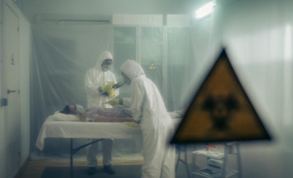 Two people attending to a woman with a virus lying on a stretcher in a field hospital with bio hazard symbol in the foreground