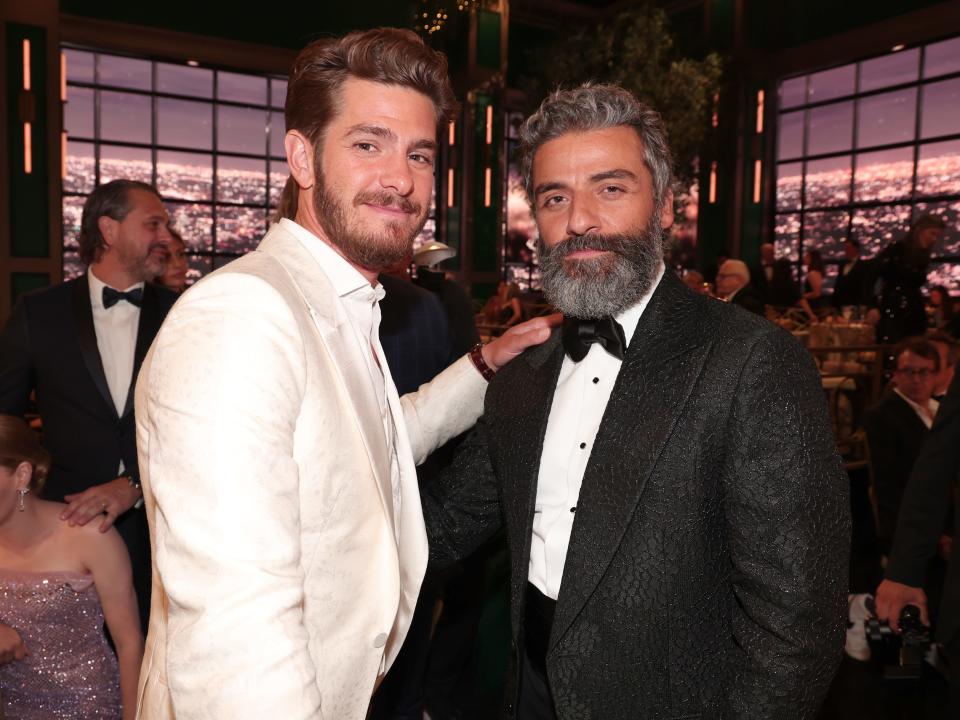 andrew garfield in a white suit with his hand on oscar isaac, who is wearing a black tuxedo,'s shoulder