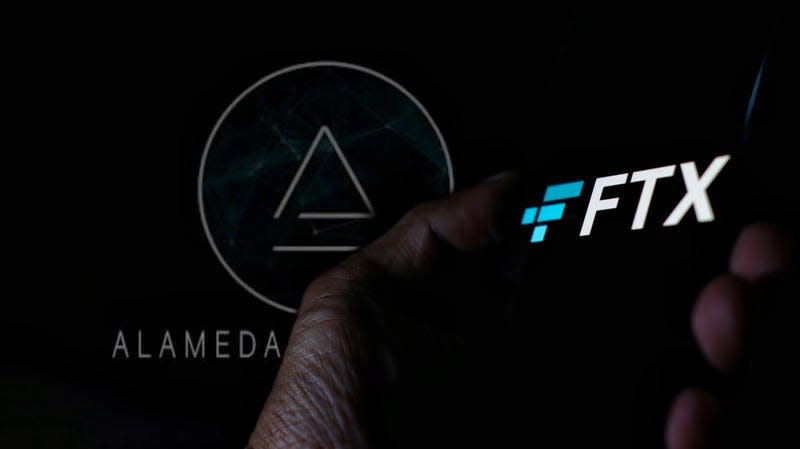 The Alameda Research symbol behind a hand holding a phone with the FTX logo.