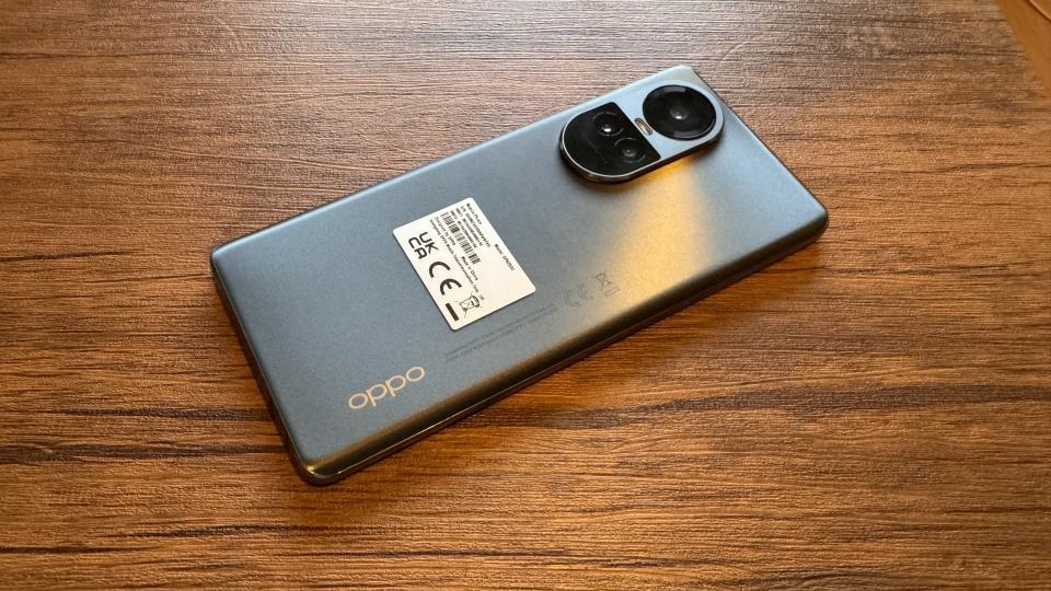 A silvery grey Oppo Reno10 camera phone sitting on a wooden table