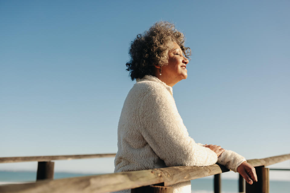 A curly-haired man in a fluffy jacket leans against a wooden railing, eyes closed, enjoying the view.