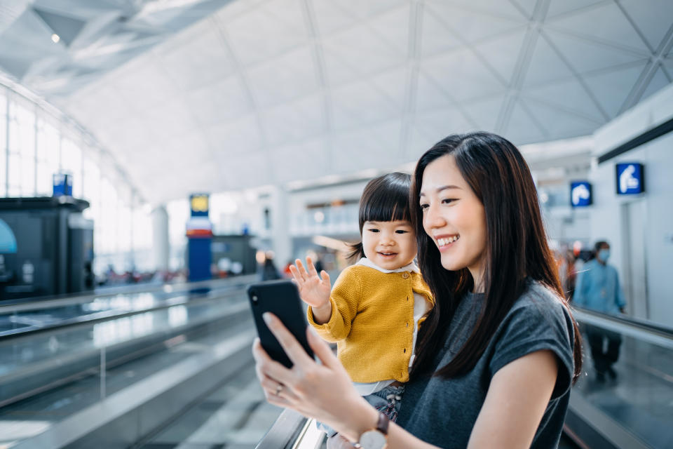 Young Asian mother and cute little daughter having video call with their family on smartphone and smiling joyfully at airport terminal while waiting for departure. Family travel and vacation. Embark on a new journey with the family