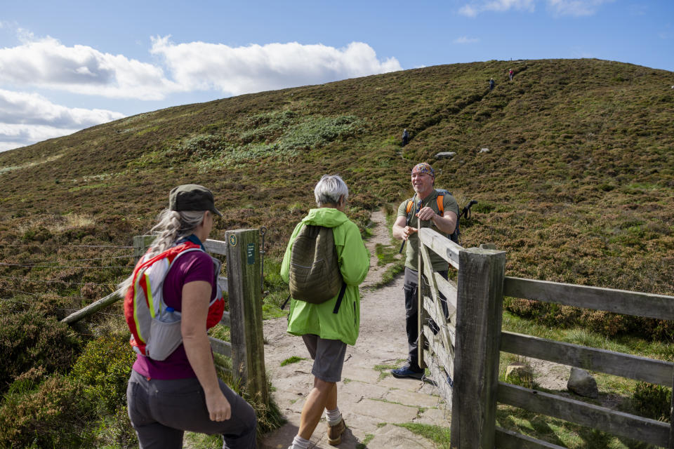 A man and woman with their mother wearing backpacks on a hike surrounded by nature in Rothbury, Northumberland. The mother and daughter are walking through a gate that the son is holding open for them.