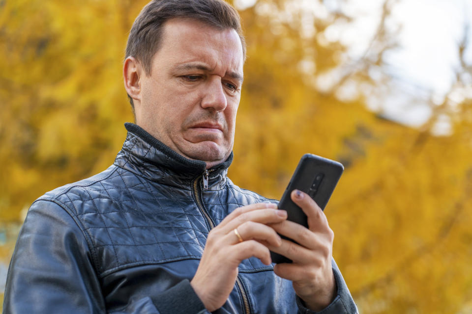 Man in a leather jacket looking at his phone with a confused expression, surrounded by autumn trees