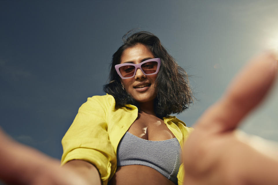 Person in sunglasses and casual summer wear looks down at the camera with arms outspread against a clear sky background