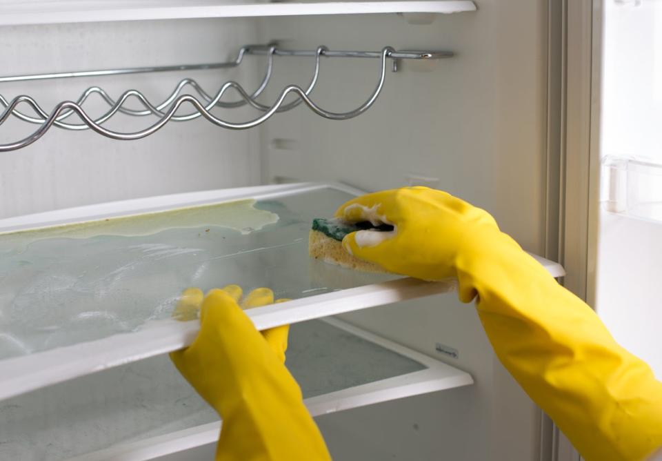 Person wearing yellow rubber gloves scrubbing refrigerator shelves with a sponge.