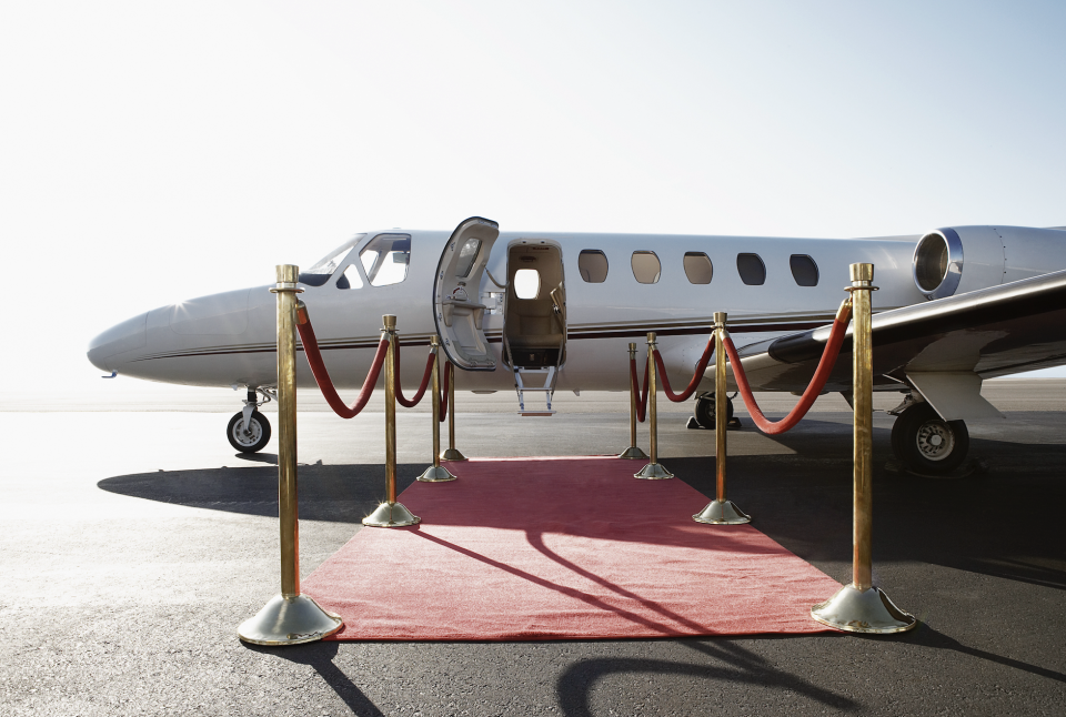 Private jet with steps down, connected to red carpet pathway framed by gold stanchions. No one is visible in the image