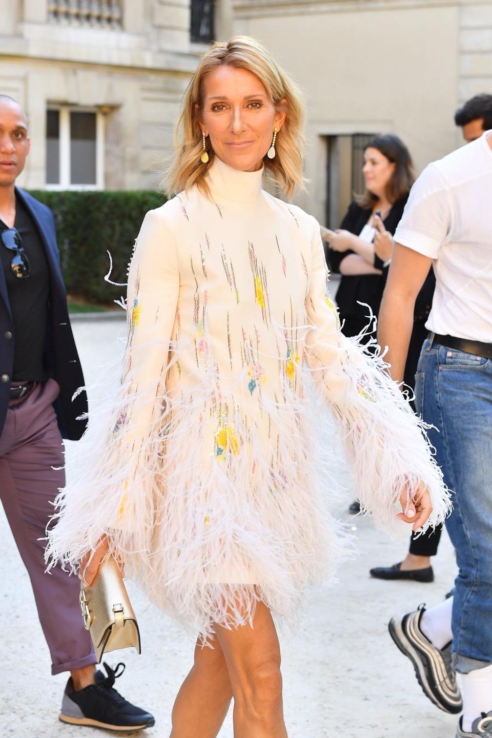 Celine in a turtleneck long-sleeve minidress. The cream dress has feathers and colorful embelishments.