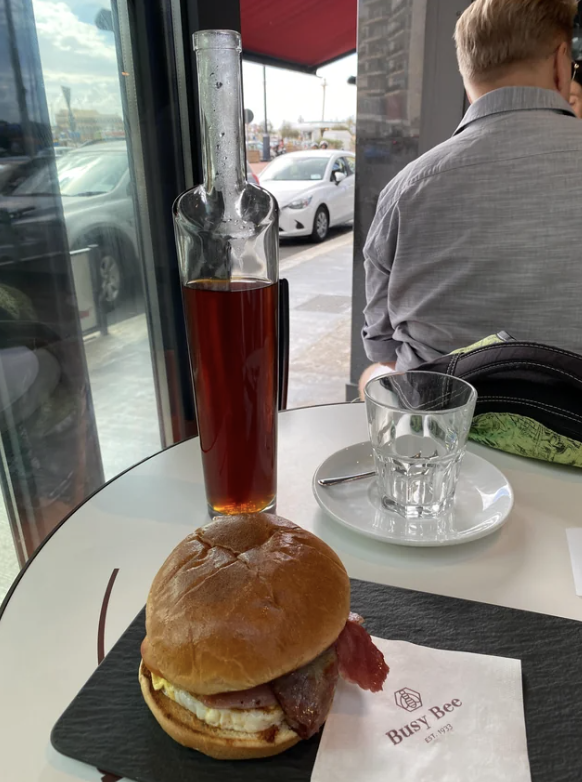 A bacon and egg burger on a plate is placed on a table with a glass bottle of sauce and an empty glass beside it. A napkin with "Busy Bee" logo is also on the table