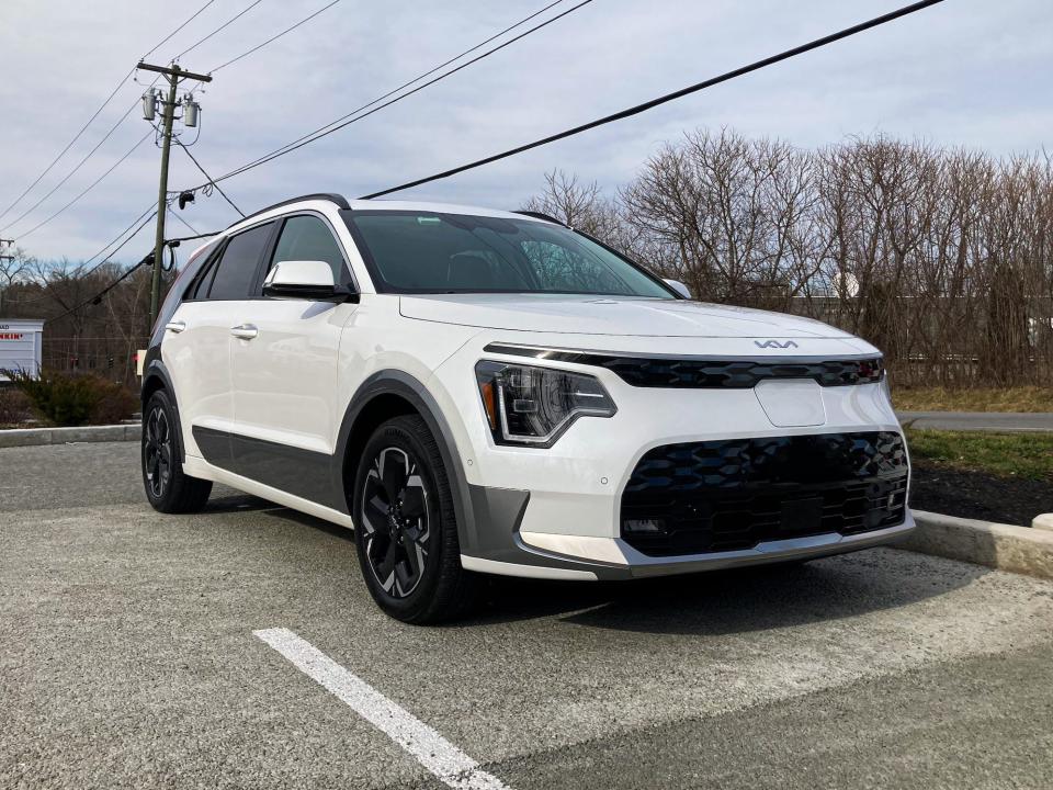 A white Kia Niro EV electric SUV is seen in a parking lot against an overcast sky, with a Tesla charging station in the foreground.