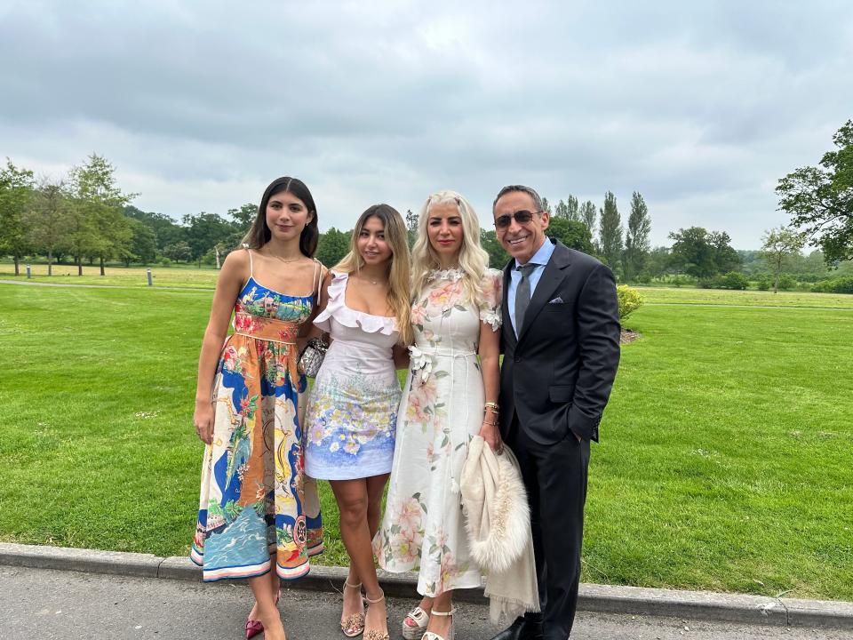 Payam Zamani with his wife and two daughters smiling and standing outside in front of a field of grass. He is wearing a suit and they are wearing dresses.