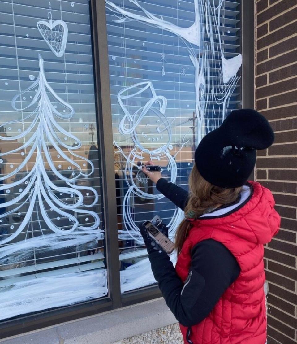 A person uses white paint to sketch out a mural design on a window.