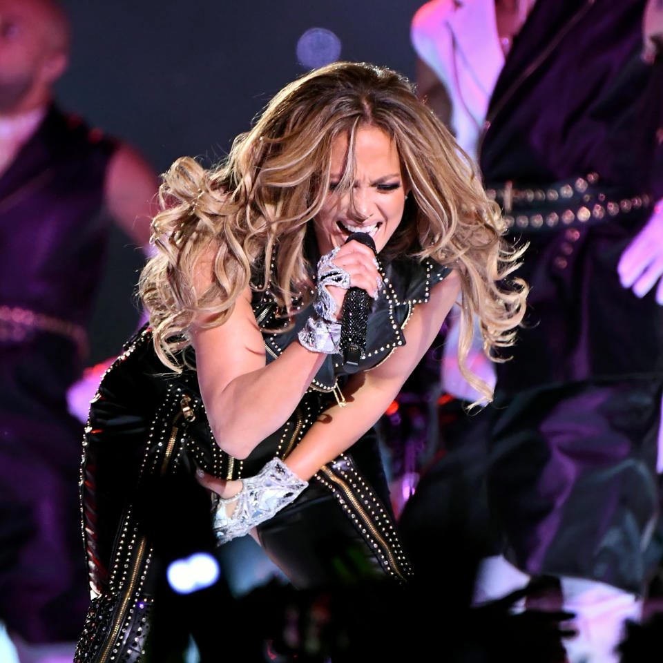 Jennifer Lopez performing energetically on stage, wearing a black studded jumpsuit with metallic gloves, singing into a microphone. Backup dancers in the background