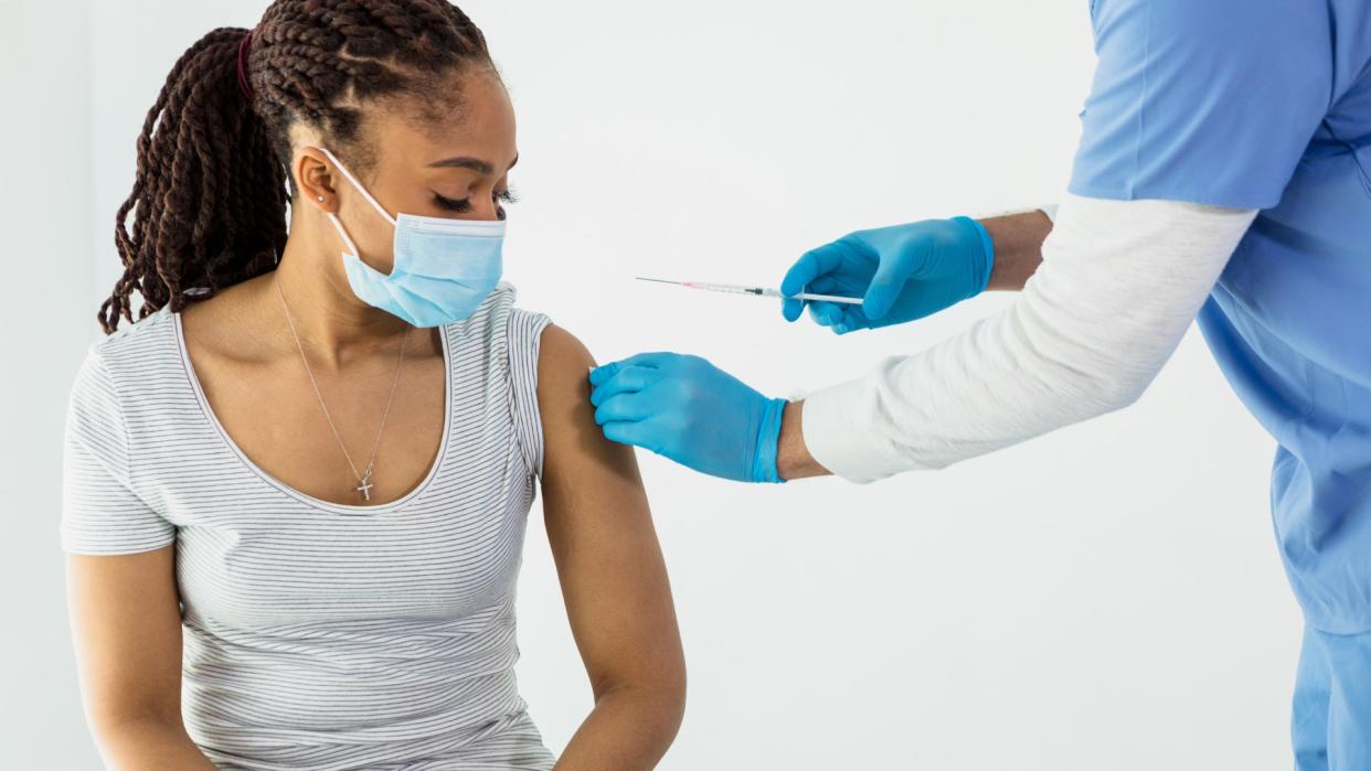  Person wearing a blue face mask sits and watches a nurse who is preparing their arm for vaccination. 