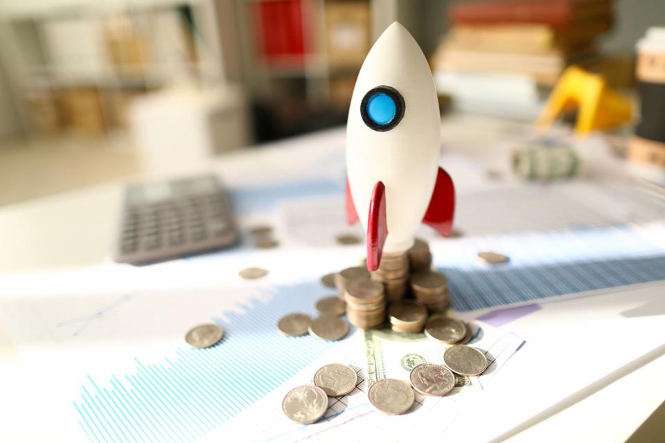 A toy rocket set for launch atop messy stacks of coins and paperwork displaying financial data. 
