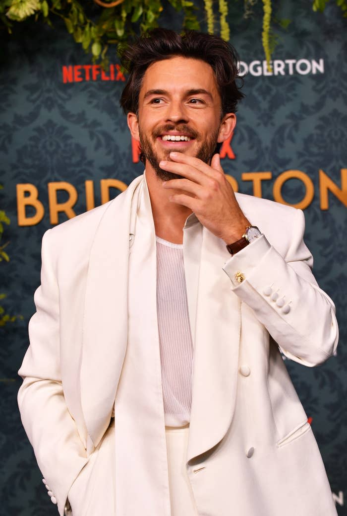 Jonathan Bailey on the Bridgerton red carpet, smiling in a white suit with a relaxed, chic style. Netflix signage is visible in the background