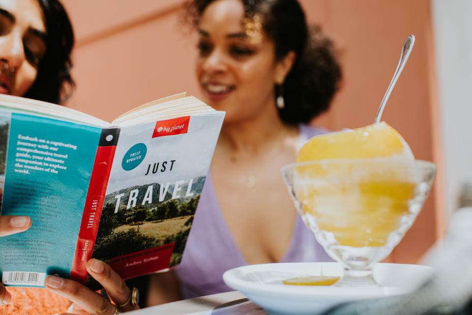 Woman reading a book titled "Just Travel" next to a bowl of dessert on a table