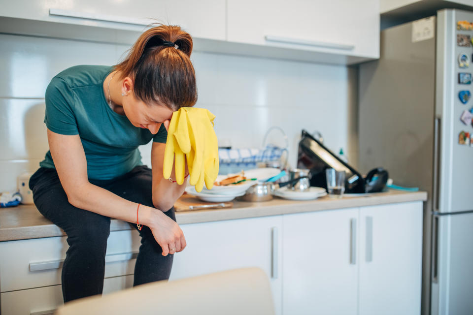 Young housewife is sitting on the kitchen counter, holding protective gloves, tired from washing dishes.