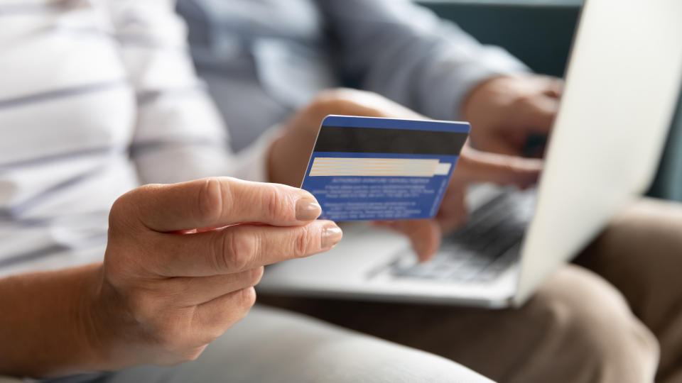 Close wife fingers holding plastic credit card on background computer on husband lap, old couple booking hotel using on-line app service and website, make online payment, shopping via internet concept