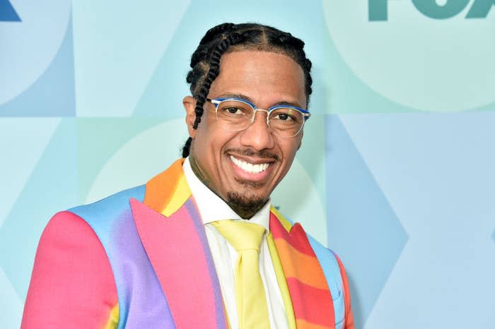 Nick Cannon is smiling, wearing a colorful, patterned suit with a shirt and a brightly-colored tie, and glasses
