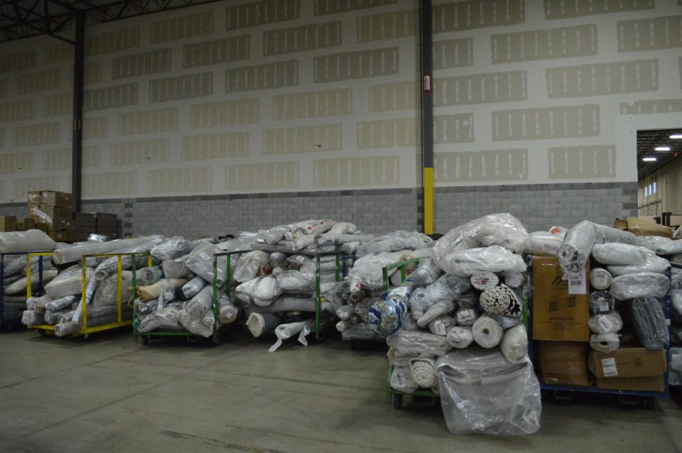 Dozens of rolled up rugs sit on cars in a grey warehouse.