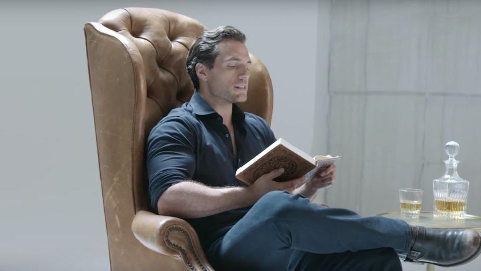 Henry Cavill sitting in a brown leather chair reading a book in The Witcher series