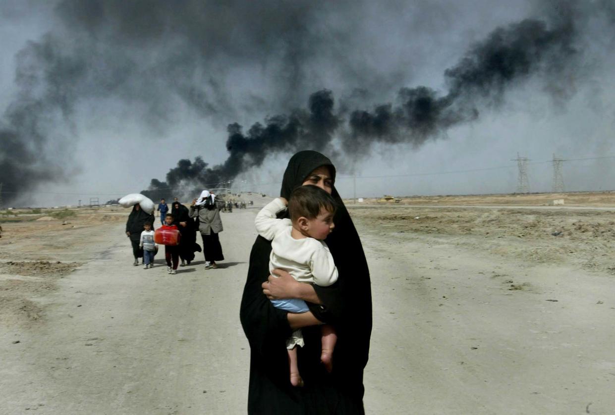 An Iraqi woman wearing a black head covering that swathes her body carries her young child, with curlicues of black smoke rising across the dirt road behind her.