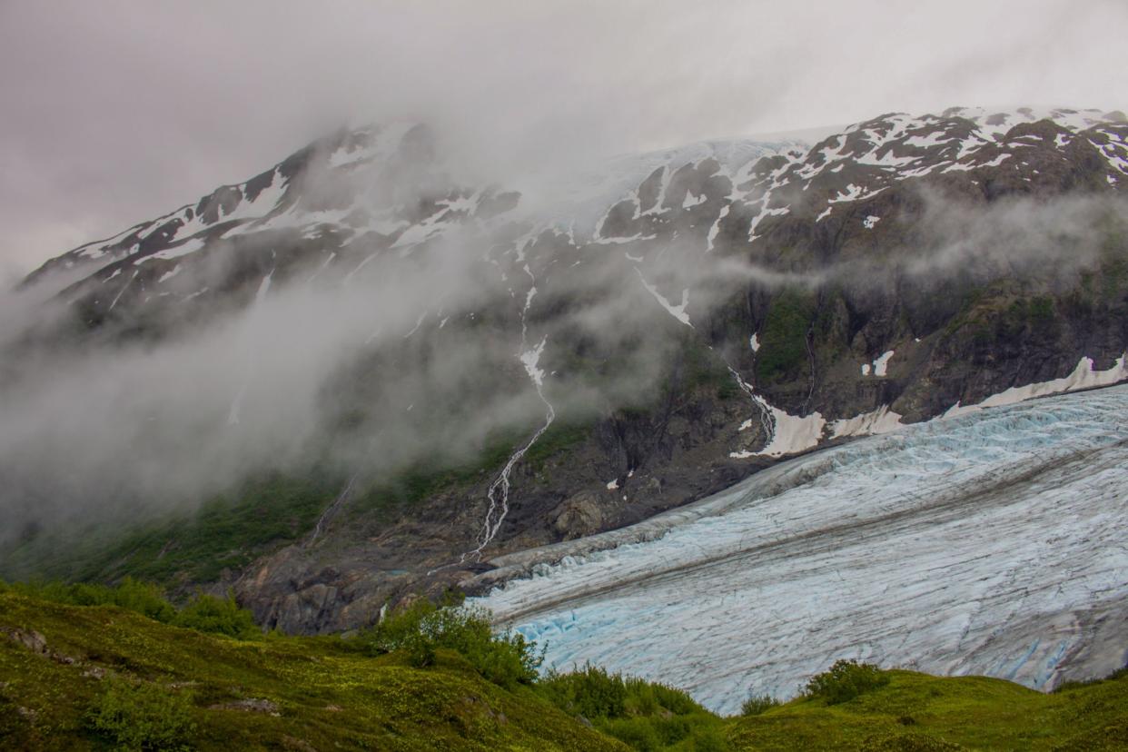 Blue and white glacier is between grassy green field with trail and snow-capped mountains covered in fog.