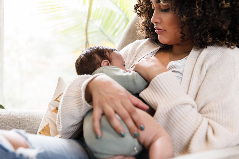 A person with curly hair breastfeeds a baby while sitting on a couch. They appear relaxed and nurturing in a cozy setting