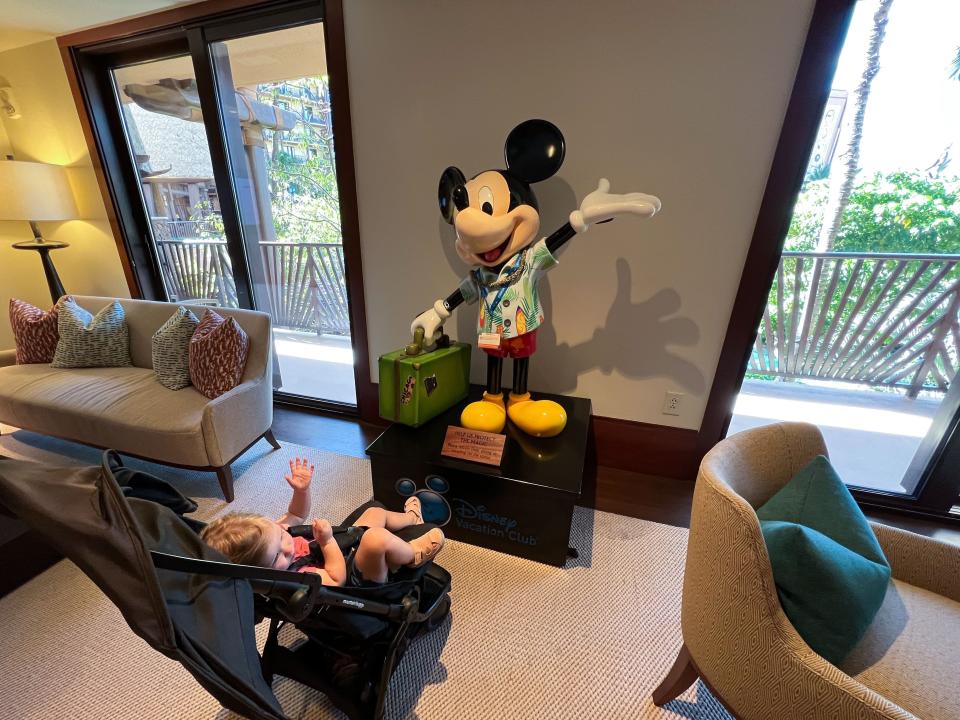 A baby in a stroller looking at a Mickey statue holding a suitcase and wearing a Hawaiian shirt.