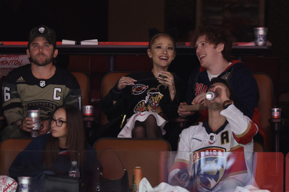 Ariana Grande and friends watch a hockey game from the audience. Ariana is smiling and holding a snack. One friend is talking to her