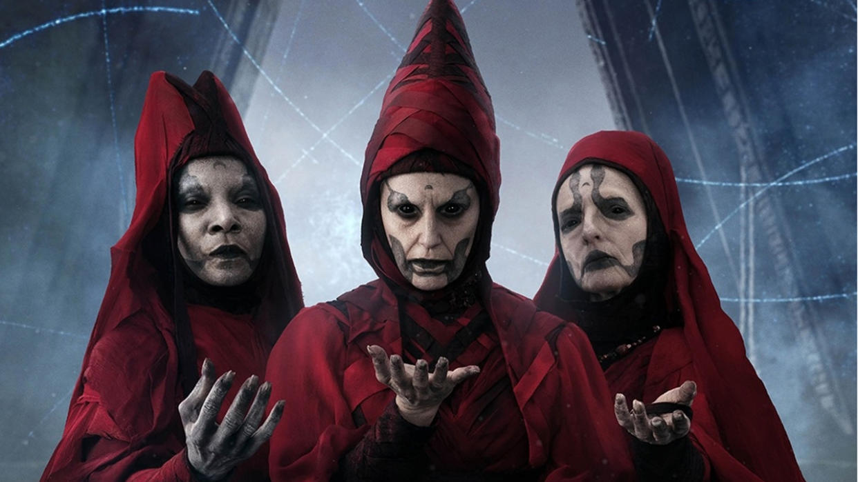  Three menacing women wearing dark red robes with hoods. They have pale white faces with black markings. They each have one hand raised, palm up. Image of some of the Witches of Dathomir from Star Wars. 