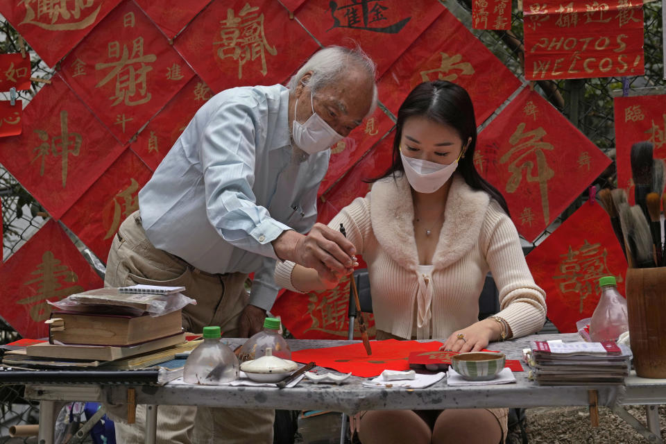 Dereck Kwok, 83-year-old calligraphy artist, left, teaches his customer Hayley Au to write "Fai Chun," traditional decorations with Chinese calligraphy in Hong Kong on Jan. 26, 2022. In the runup to the Lunar New Year, calligraphers set up on the streets of Hong Kong to write ink-brush phrases on traditional red paper banners for homes and offices. (AP Photo/Kin Cheung)