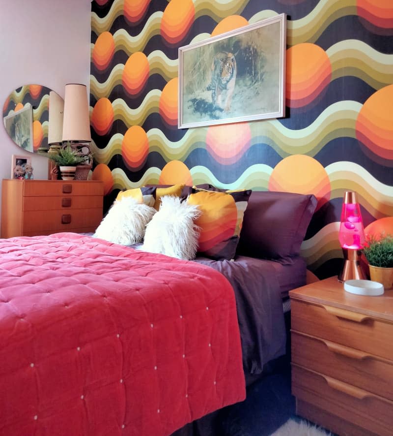 Bedroom with colorful wall graphic wallpaper. Bed next to a pink lava lamp.