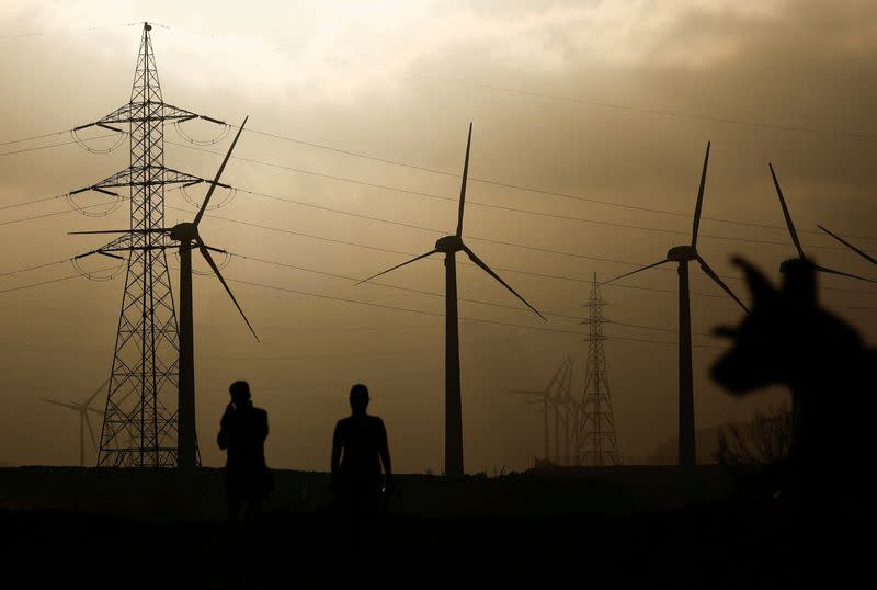 Dos personas caminan cerca de una torre eléctrica y un grupo de turbinas eólicas, en Barranco de Tirajana