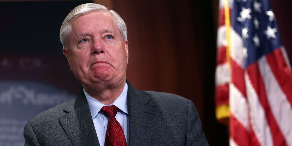 lindsey graham wearing a gray suit and red tie, frowning and looking off camera, with an american flag behind him