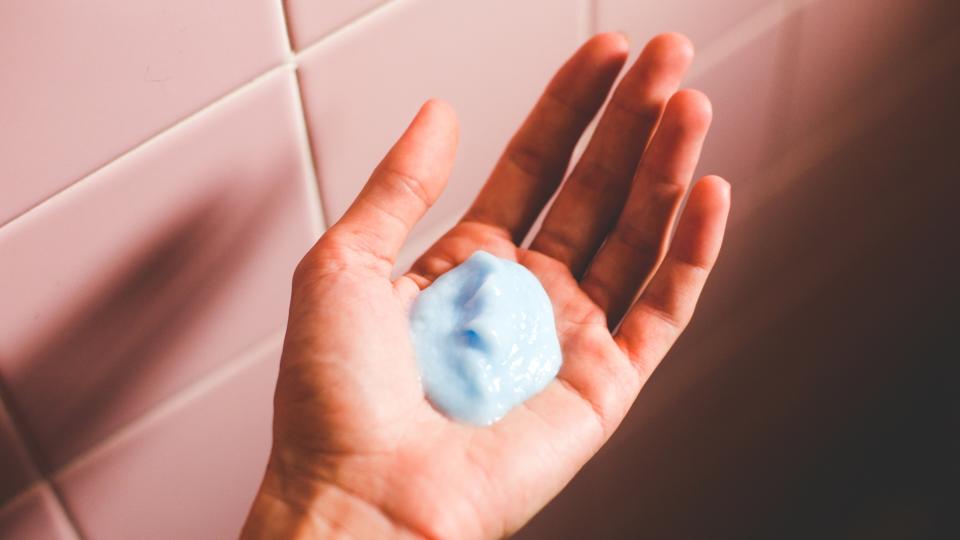 A woman's hand pictured holding a cream hair product in her palm, in front of a pink bathroom backdrop