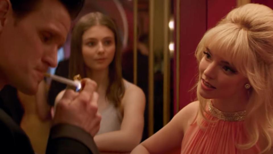 Matt Smith lights a cigarette while Anya Taylor-Joy talks to him, and Thomasin McKenzie watches from the mirror in Edgar Wright's Last Night in Soho.